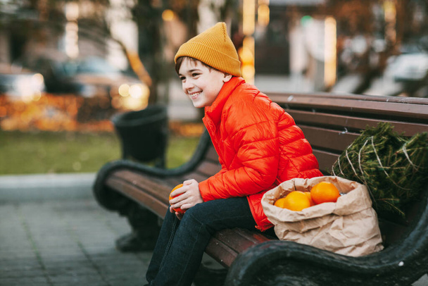 Een jongen in een fel oranje jasje en een modieuze gele hoed zit op een bankje, lacht, gooit sinaasappels in de lucht, en naast hem staat een boompje en een grote zak sinaasappels. Winkelen voor de - Foto, afbeelding