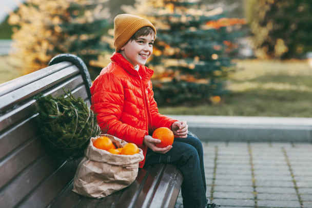 Chlapec v jasně oranžovém saku a módním žlutém klobouku sedí na lavičce, směje se, vyhazuje pomeranče do vzduchu a vedle něj je malý stromek a velký pytel pomerančů. Nakupování pro - Fotografie, Obrázek