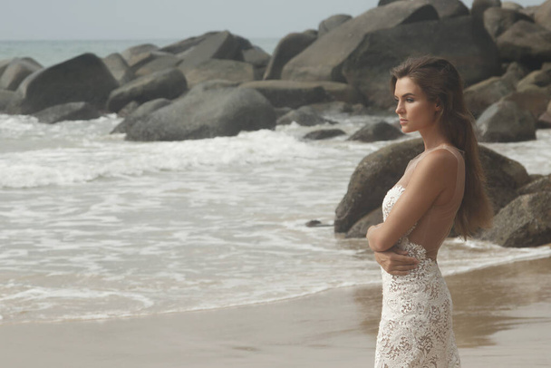Young woman wearing beautiful white dress is posing on the rocky beach - Фото, зображення