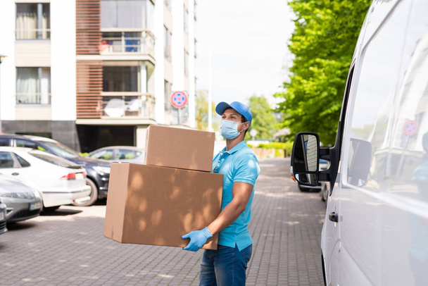 Delivery man wearing prevention mask and gloves is holding stack of the cardboard boxes near his van - Фото, изображение