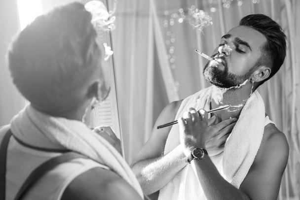Handsome man looking in the mirror, smoking a cigarette and shaving his beard with a straight razor - Foto, immagini