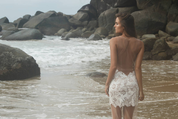Young woman wearing beautiful white dress is posing on the rocky beach - Фото, изображение
