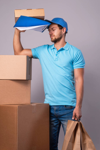 Vermoeide bezorger tijdens het werk met een pakje in een kartonnen doos over grijze achtergrond - Foto, afbeelding
