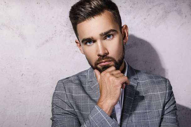 Handsome bearded man wearing gray checkered suit is posing against concrete wall - Фото, зображення