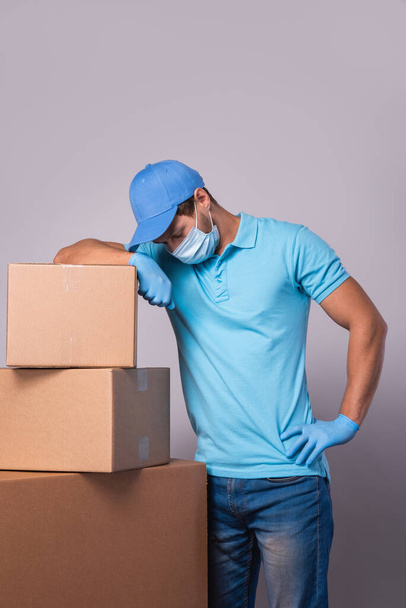 Young delivery man is wearing prevention mask and gloves with a cardboard boxes isolated on gray background - Фото, изображение