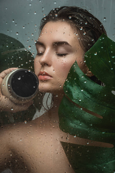 Portrait of beautiful woman with a tropical leaf captured through wet glass - Fotó, kép