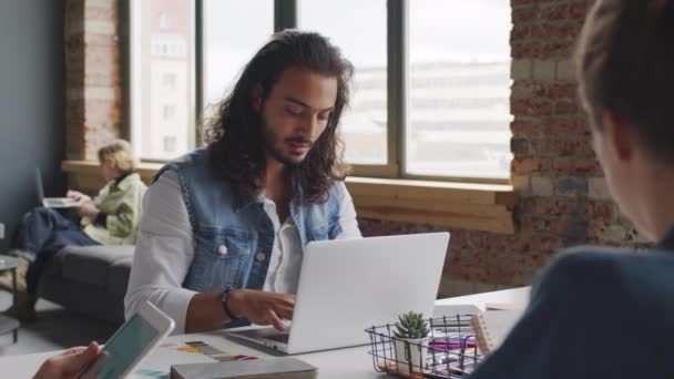PAN slowmo młodego mężczyzny start-up pracownika firmy z włosami na ramionach siedzi przy stole w sali konferencyjnej nowoczesnego biura i wpisując na laptopie, jak jego koledzy pracują obok niego - Materiał filmowy, wideo