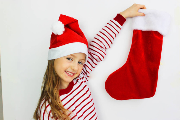 Beautiful girl red sweater holding a Christmas sock and waiting for gifts from Santa . Christmas and New year gift. Happy small girl in Santa s hat has a Christmas. - Fotoğraf, Görsel