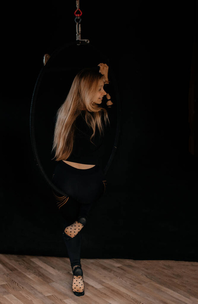 A graceful young woman performs an acrobatic element on an air ring in a dark room. Gymnastic ring. Gymnastics and sports concept. - Zdjęcie, obraz
