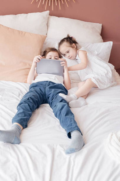A little boy and a girl with special needs lie on a bed in a room playing on a tablet. Boy in a white shirt with a tablet. The concept of caring for children with special needs.  Copy space. Toning. - Zdjęcie, obraz