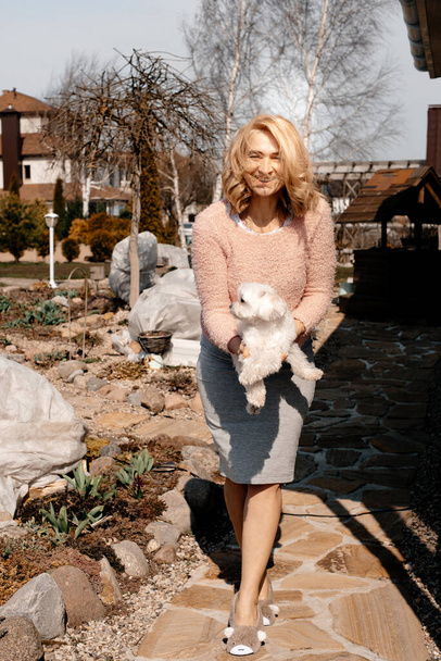 A blonde girl in a gray skirt holds a small dog in her arms, against the background of the park. Summer sunny day. Small purebred white dog. Pet. - Foto, imagen