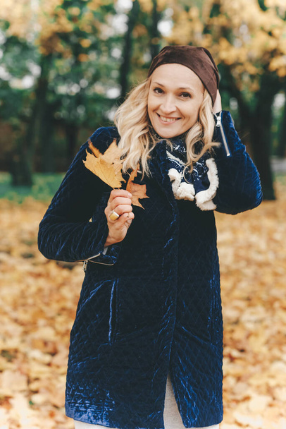 Happy girl with blond hair in a coat walks in the autumn park. Girl with leaves in her hands. Yellow autumn foliage fallen to the ground. - Foto, imagen