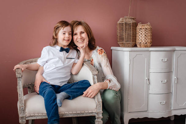 Mom and a little boy with Down Syndrome are sitting in a room on an armchair and smiling.Happy family mom and son. The concept of caring for children with special needs.  Copy space. Toning. - Fotografie, Obrázek