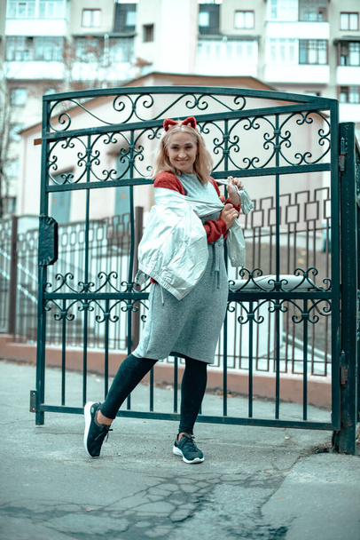 A sweet women with blond hair in a gray dress and with a hoop ears near a black iron fence. The woman on the other side of the fence. The girl walks around the city. - Foto, Bild