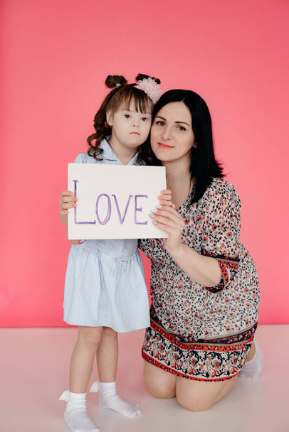 A girl with Down Syndrome and her mother are holding a sign that says love. Pink plain background in the background. Disabled care concept.  Copy space. Toning. - Photo, image
