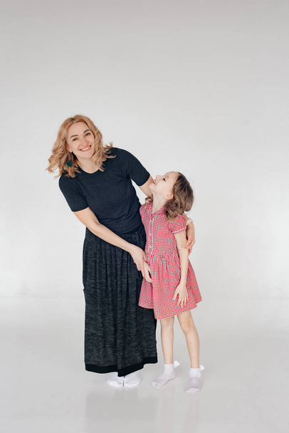 Mom in a black long dress hugs her daughter smiling, in the background a single-ton white background. The concept of style and fashion. Spend time together taking pictures.  Copy space. Toning. - 写真・画像