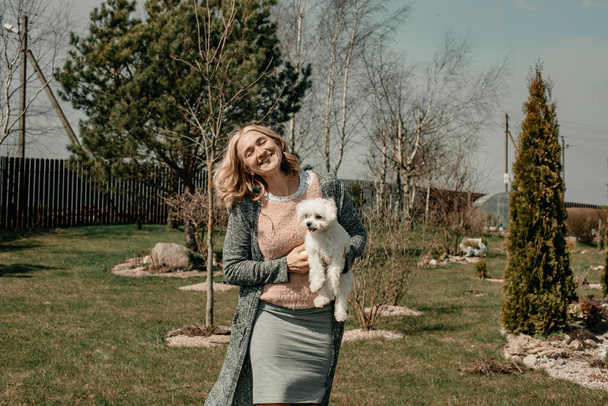 An attractive young blonde girl holds a small puppy in her arms on a sunny summer day against the backdrop of trees. Small purebred white dog. Pet. The concept of happiness and positivity. - Фото, изображение