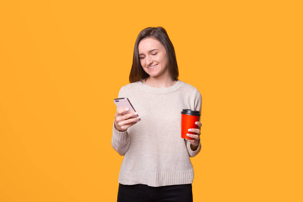 Jeune femme lit ou envoie des SMS sur son téléphone tout en tenant une tasse à emporter . - Photo, image