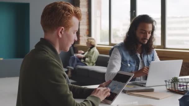 PAN slowmo των δημιουργικών νέων start-up εργαζομένων της εταιρείας κάθεται μαζί σε ευρύχωρη αίθουσα συνεδριάσεων του σύγχρονου γραφείου και εργάζονται σε gadgets. Άνθρωπος με κόκκινα μαλλιά κοιτάζοντας τις ιδέες interface app στο tablet - Πλάνα, βίντεο