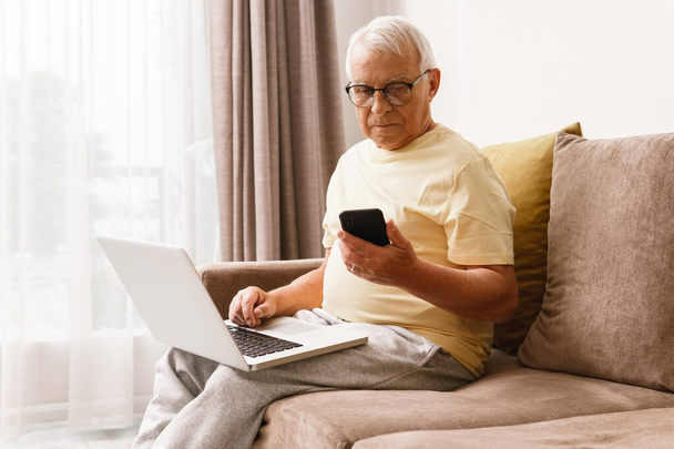 Senior man  wearing eyeglasses is using laptop computer and smartphone at home - Photo, Image