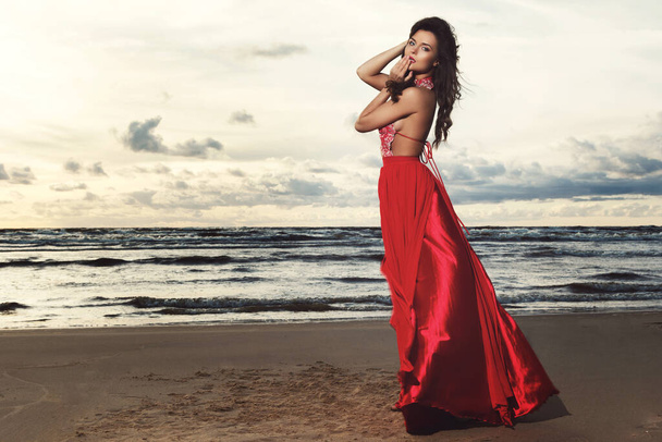 Stunning woman wearing beautiful red dress on the beach during sunset time - Zdjęcie, obraz