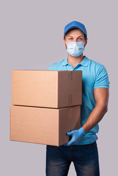 Young delivery man is wearing prevention mask and gloves with a cardboard boxes isolated on gray background - Fotó, kép