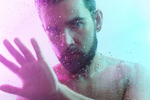 Portrait of handsome young man through wet glass in neon light - Фото, изображение