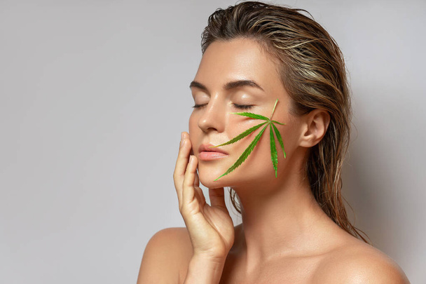 CBD cosmetics concept. Beautiful woman with a cannabis leaf on gray background - Фото, изображение