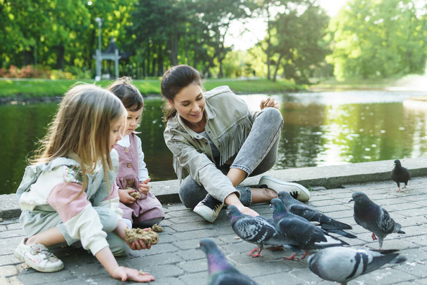 Young mother and her cute little daughters feeding birds in city park - Photo, Image