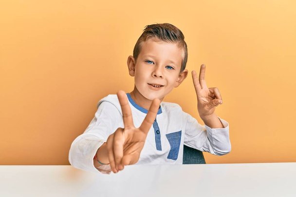 Entzückendes kaukasisches Kind in lässiger Kleidung, das auf dem Tisch sitzt und lächelnd in die Kamera blickt, die Finger beim Siegeszeichen zeigt. Nummer zwei.  - Foto, Bild