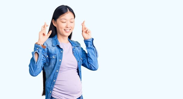 Joven hermosa mujer china embarazada esperando bebé gesto dedo cruzado sonriendo con esperanza y los ojos cerrados. suerte y concepto supersticioso.  - Foto, Imagen