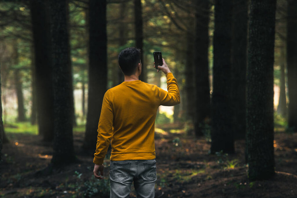 person with yellow jacket and gray jeans taking photos with mobile in the forest - Фото, изображение