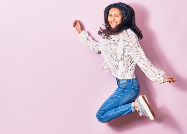 Jonge mooie latijnse vrouw draagt casual kleding glimlachend gelukkig. Springen met een glimlach op het gezicht over geïsoleerde roze achtergrond - Foto, afbeelding
