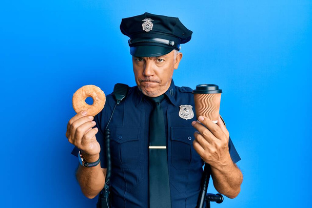 Guapo maduro policía de mediana edad comiendo donut y bebiendo café escéptico y nervioso, frunciendo el ceño molesto debido a un problema. persona negativa.  - Foto, Imagen