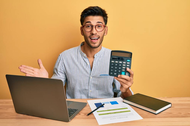 młody przystojny mężczyzna pokazując kalkulator w biuro  - Zdjęcie, obraz