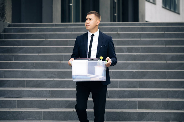 Male office worker in depression with box of personal stuff. Left without money. Businessman lost job. Fired man walking outdoor. Depressed jobless person. Unemployment concept - Zdjęcie, obraz