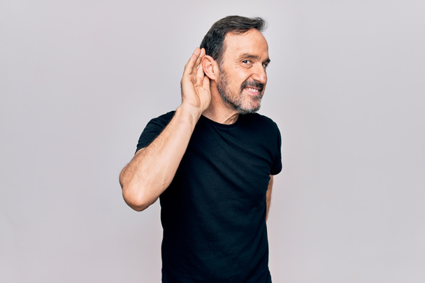 Middle age man wearing casual black t-shirt standing over isolated white background smiling with hand over ear listening and hearing to rumor or gossip. Deafness concept. - Photo, Image