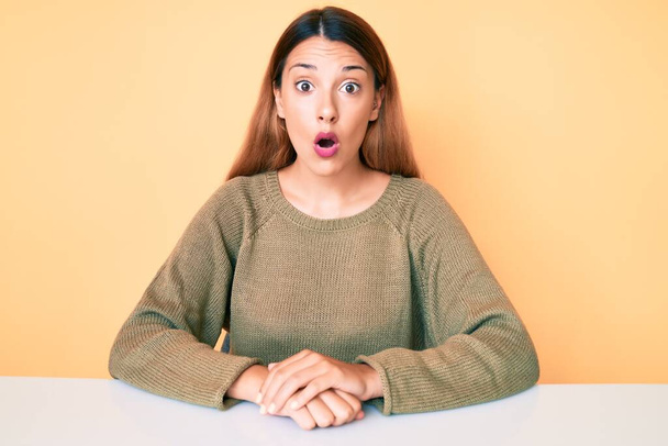 Jeune femme brune portant des vêtements décontractés assis sur la table effrayé et étonné avec la bouche ouverte pour la surprise, visage incrédule  - Photo, image