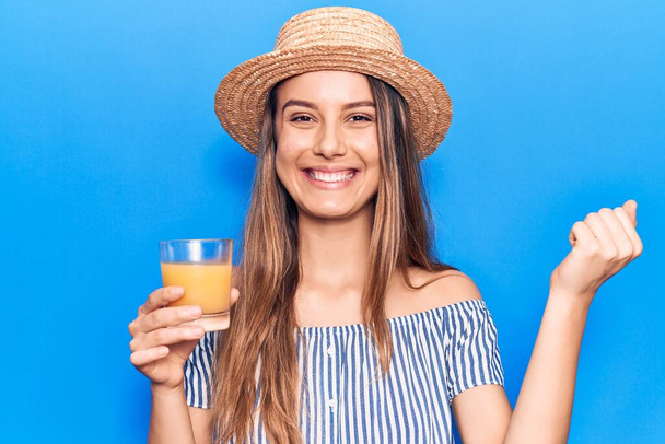 Joven hermosa chica con sombrero de verano beber vaso de jugo de naranja gritando orgulloso, celebrando la victoria y el éxito muy emocionado con el brazo levantado  - Foto, imagen