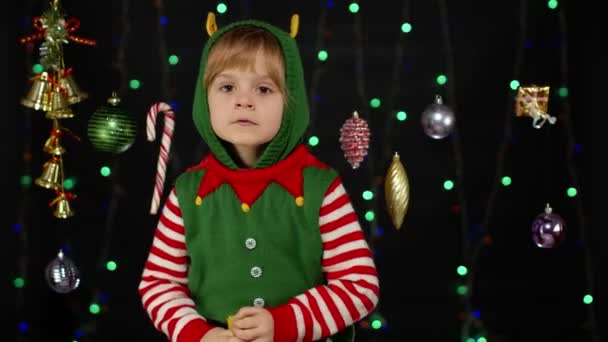Kid girl in Christmas elf Santa Claus helper costume looks at camera makes big eyes covering mouth - Footage, Video