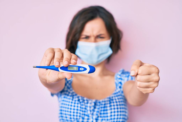 Young beautiful woman wearing medical mask holding holding thermometer annoyed and frustrated shouting with anger, yelling crazy with anger and hand raised  - Photo, Image