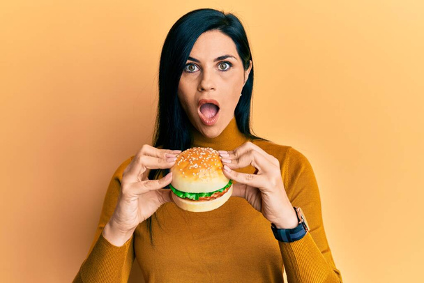 Young caucasian woman eating a tasty classic burger afraid and shocked with surprise and amazed expression, fear and excited face.  - Photo, Image