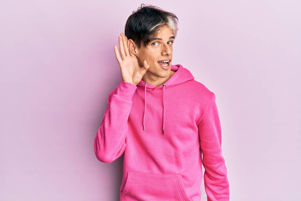 Junger hispanischer Mann in lässigem rosa Sweatshirt, der lächelnd mit der Hand über dem Ohr Gerüchten oder Klatsch lauscht und zuhört. Konzept für Gehörlosigkeit.  - Foto, Bild