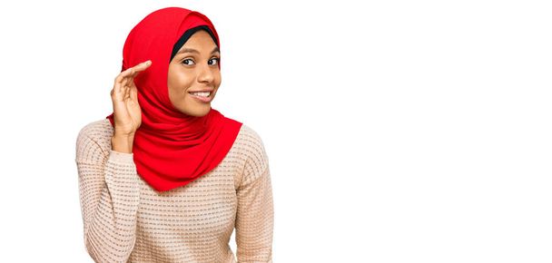 Молода афроамериканська жінка, одягнена в традиційний ісламічний шарф хіджабу, посміхається з рукою за вухом, слухаючи слух до пліток або пліток. концепція глухоти.  - Фото, зображення
