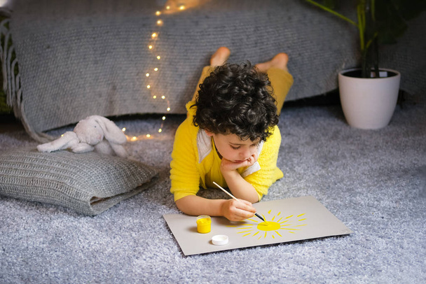 Little curly boy drawing yellow sun in gray interior. - Foto, afbeelding