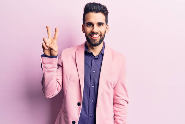 Mladý pohledný muž s vousy v elegantní bundě ukazuje a ukazuje prsty číslo dvě a zároveň se usmívá sebevědomě a šťastně.  - Fotografie, Obrázek