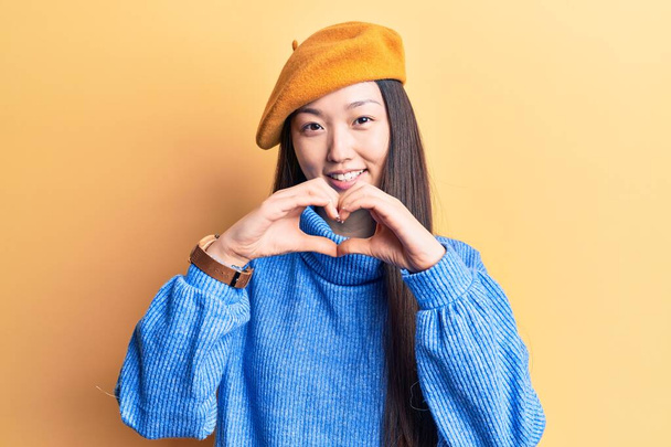 Młoda piękna Chinka nosząca sweter z golfem i francuski beret uśmiechająca się w miłości robiąc ręcznie kształt symbolu serca. romantyczna koncepcja.  - Zdjęcie, obraz