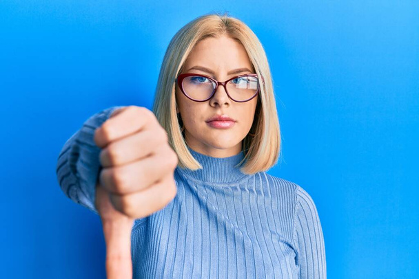 Young blonde woman wearing casual clothes and glasses looking unhappy and angry showing rejection and negative with thumbs down gesture. bad expression.  - Photo, Image