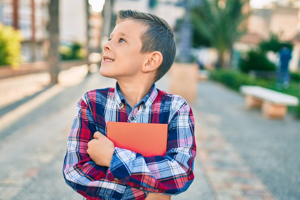 Adorável estudante caucasiano menino sorrindo feliz segurando livro de pé na cidade. - Foto, Imagem