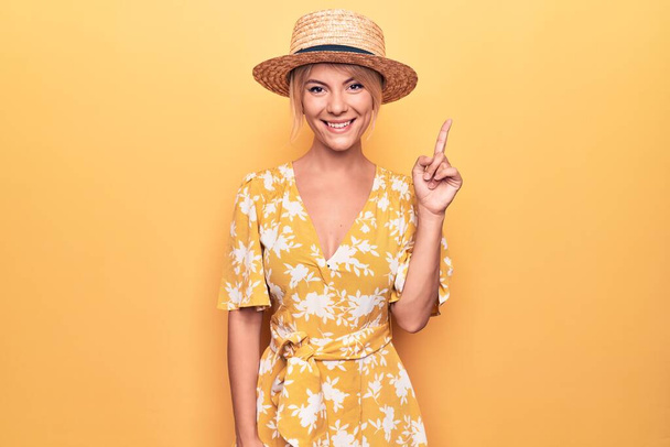 Красивая блондинка на отдыхе в летней шляпе и платье на желтом фоне, показывая и показывая пальцем номер один, улыбаясь уверенно и счастливо. - Фото, изображение